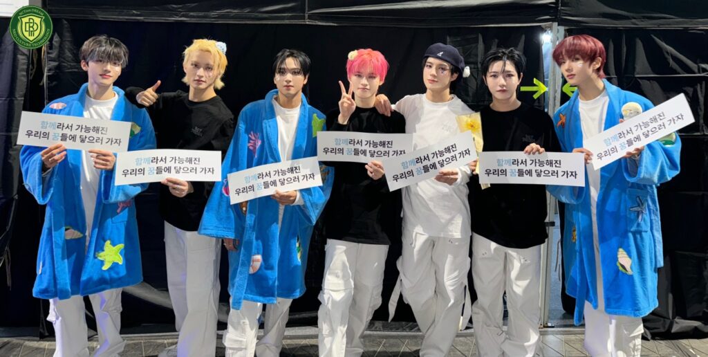 NCT DREAM members backstage holding signs after their performance, wearing blue robes and white outfits